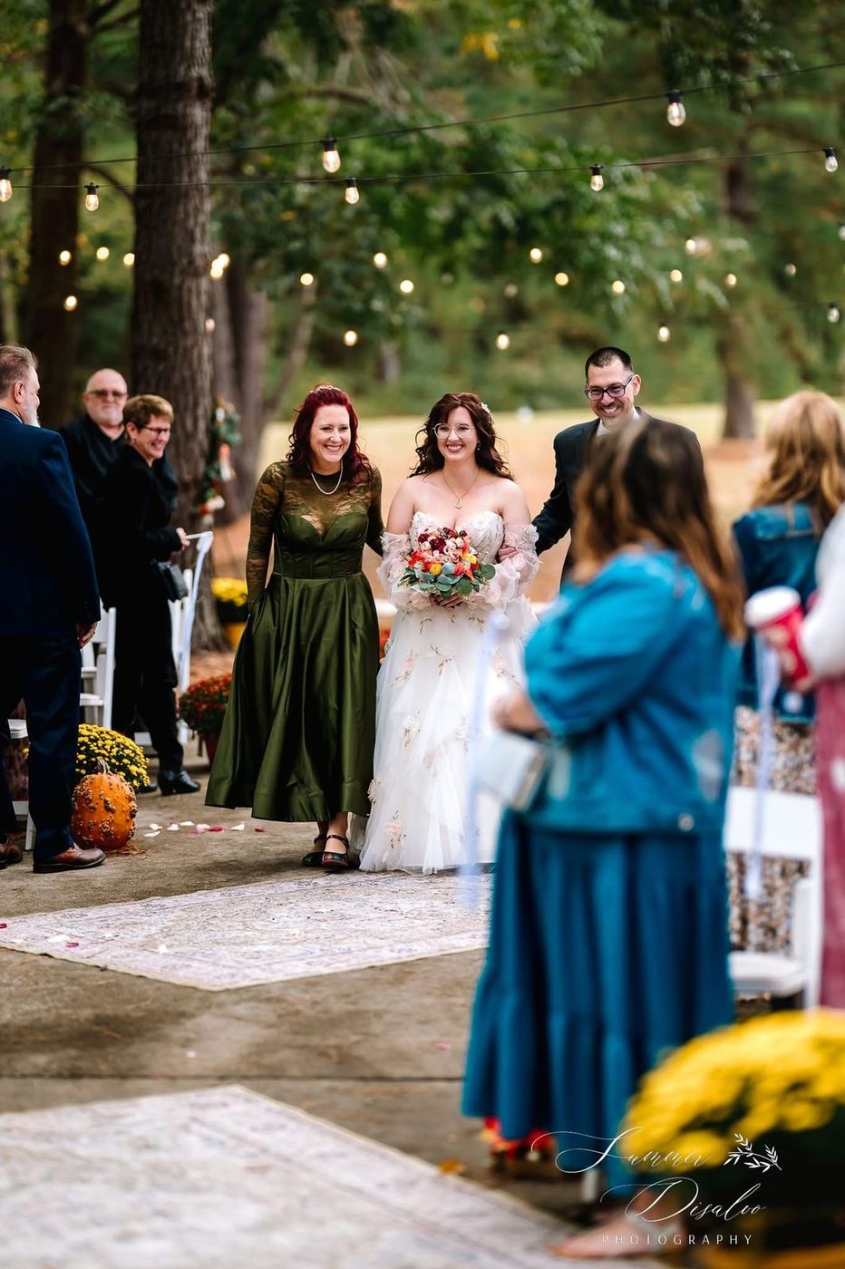 Bride walking down an outdoor aisle under string lights with her parents, past mums and pumpkins (photographer watermark shown)