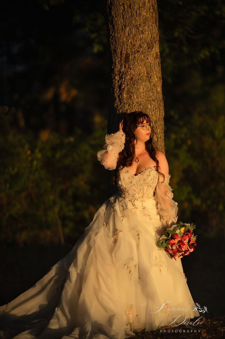 Bride in a floral tulle gown leaning against a tree in warm evening light (photographer watermark shown)