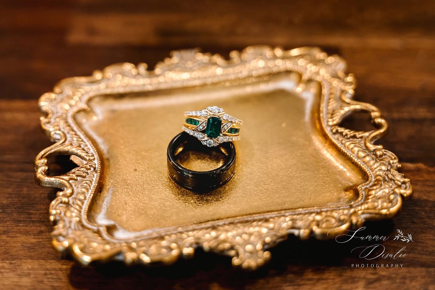 Emerald and diamond ring set with a black wedding band on an ornate gold tray (photographer watermark shown)