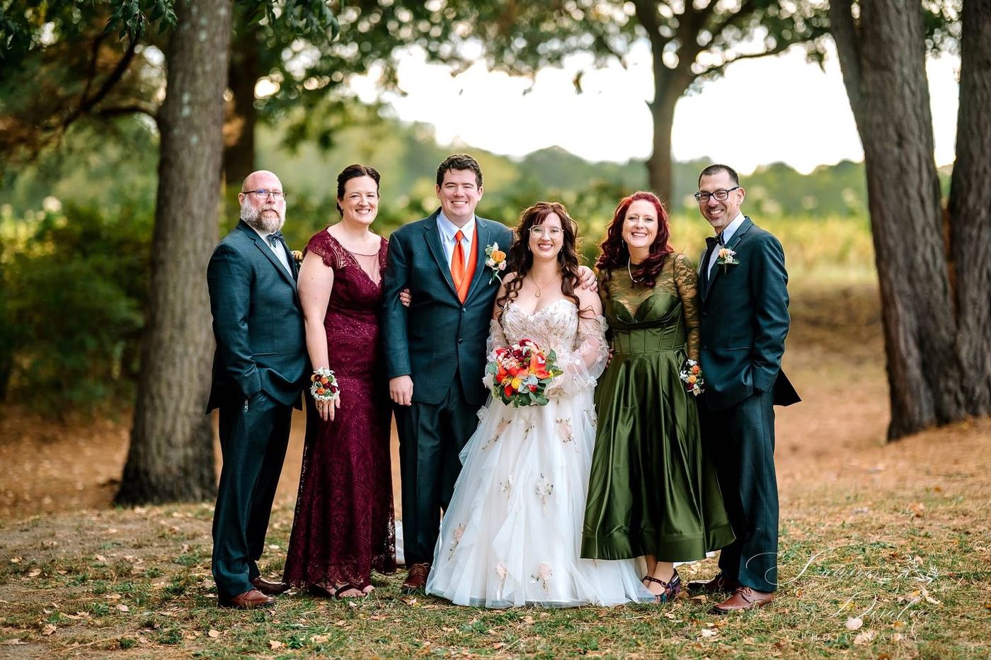 Couple with their parents in autumn tones beneath the trees (photographer watermark shown)