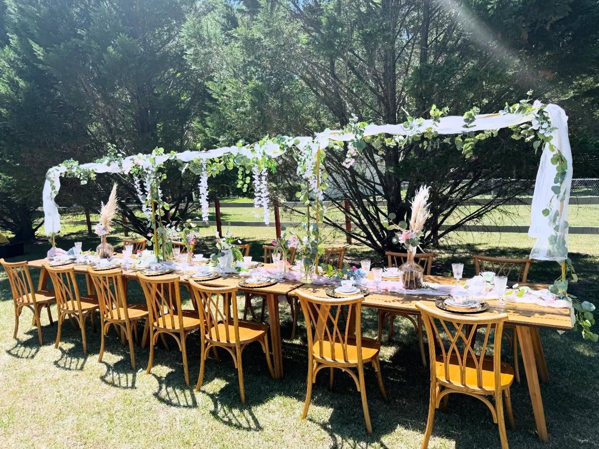 Long outdoor farm table under a draped greenery canopy with wisteria, pampas and vintage place settings