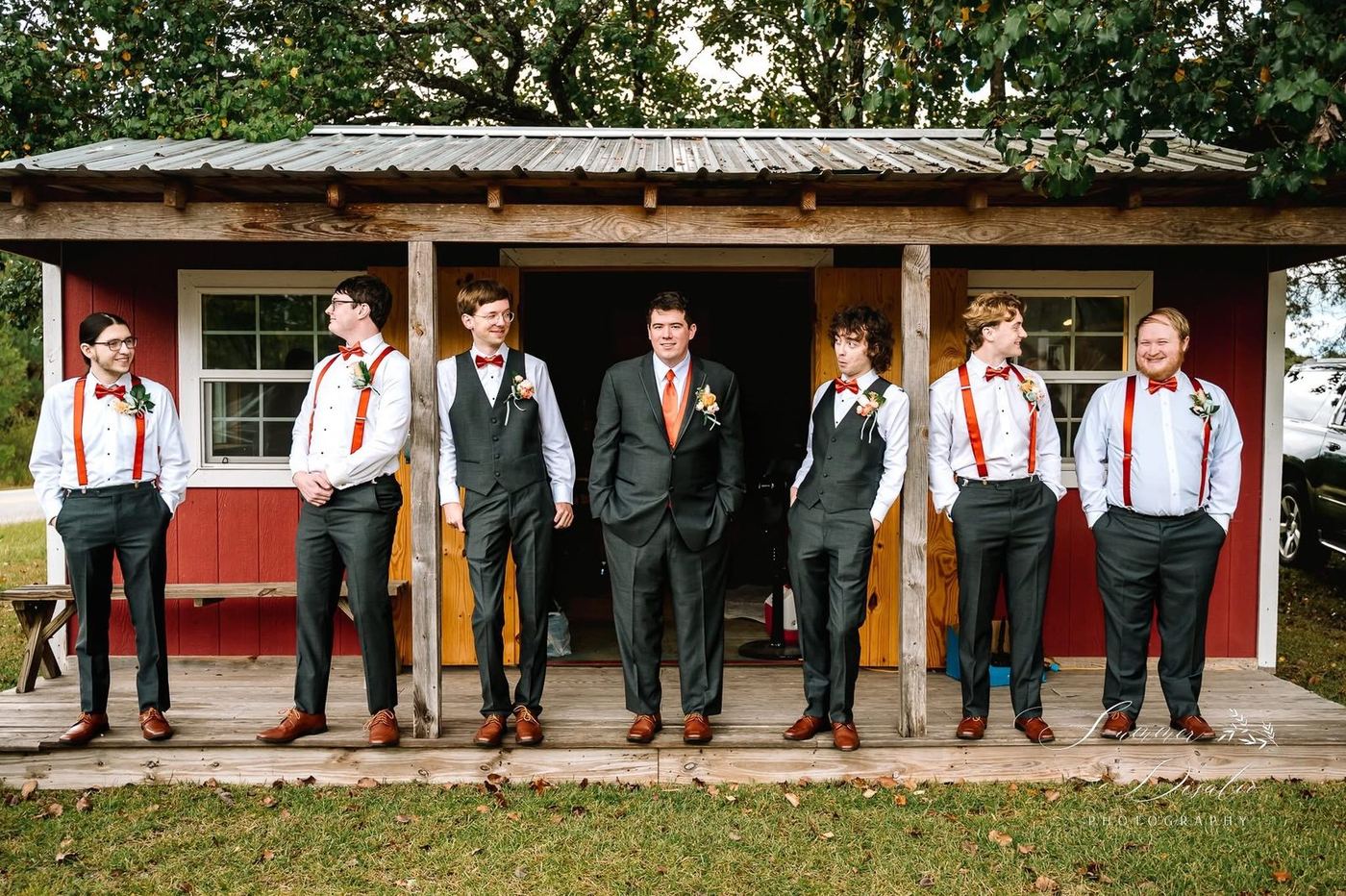 Groom and groomsmen in charcoal suits with orange bow ties and suspenders on a red cabin porch (photographer watermark shown)