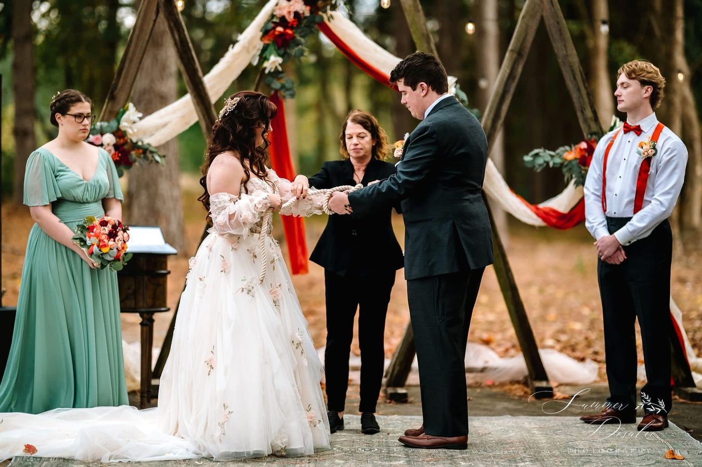 Handfasting rope ceremony beneath a draped triangle arch with a bridesmaid and groomsman (photographer watermark shown)