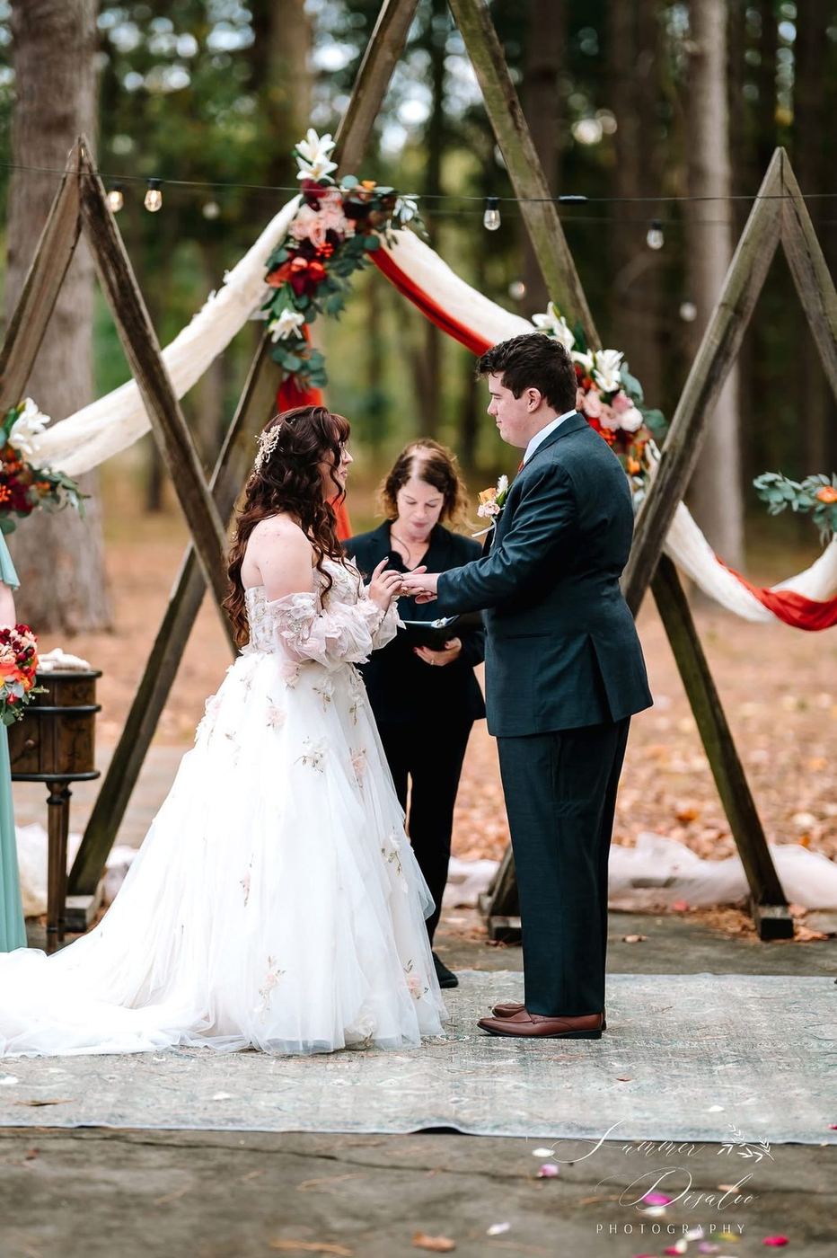 Ring exchange beneath a wooden triangle arch draped in ivory and rust fabric with autumn florals (photographer watermark shown)