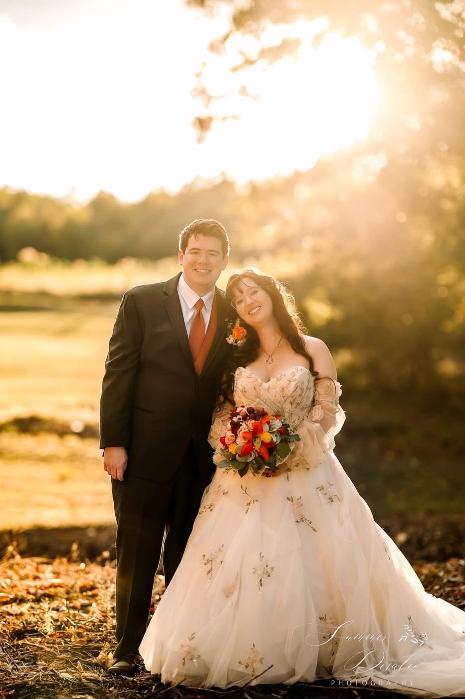 Couple smiling at sunset with an autumn bouquet of red lilies, roses and eucalyptus (photographer watermark shown)