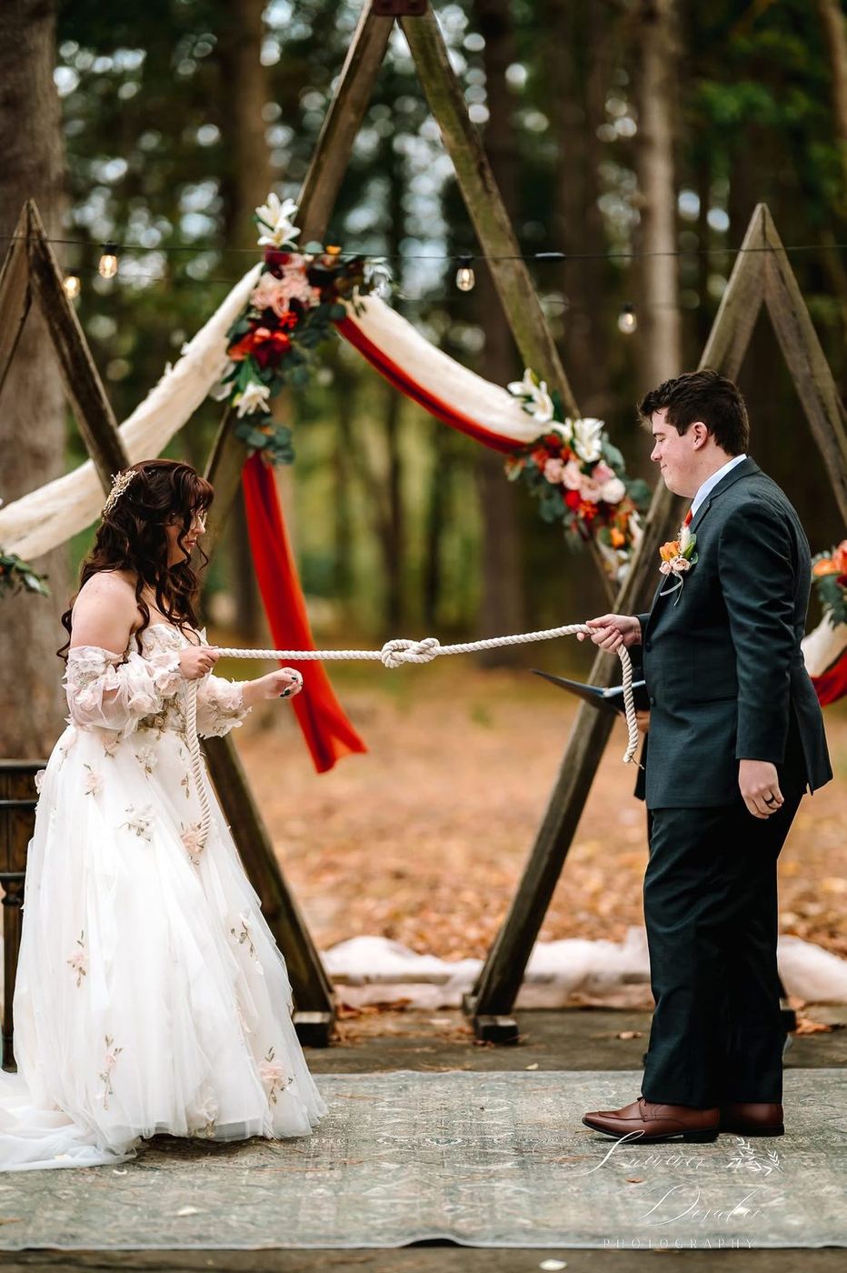 Couple tying a unity knot in front of a fabric-draped wooden arch with autumn florals (photographer watermark shown)