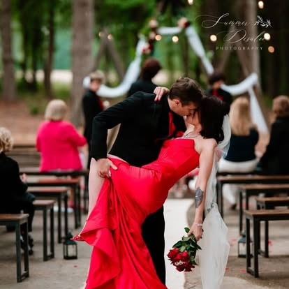 Bride in a red gown dipped for a kiss by the groom at an outdoor ceremony (photographer watermark shown)